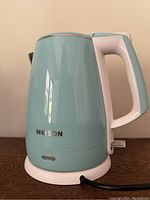Front view of Meison electric tea kettle showing light turquoise body, white handle, and brand name on the kettle.