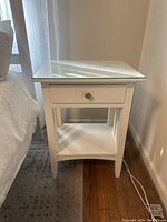 Front angle view of one white side table showing glass top, single drawer with brass knob, lower shelf, and straight legs.