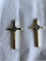 Front views of two gold-tone metal crosses with silver-tone center medallions and hanging rings placed on crumpled white paper.