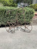 Vintage rustic wire bicycle plant stand sitting outside on concrete with greenery in background, showing wire frame and baskets clearly.