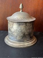 Front angle of vintage British metal tea canister showing tarnished surface and floral engravings on lid and body