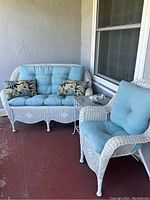 Wide view of white wicker loveseat with light blue cushions and patterned throw pillows, adjacent to matching armchair with blue cushions, on red concrete floor