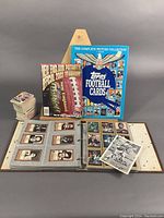 Wide shot showing New England Patriots Official 2003 Yearbook, Topps Football Cards The Complete Picture Collection book, signed O.J. Simpson photo, binder with football cards, and stack of loose cards