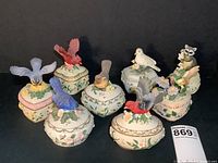 Eight Heritage House ceramic trinket music boxes with bird and raccoon figurines on heart-shaped floral bases, arranged on a black background.