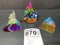 Three crystal paperweights arranged in a row on a dark surface showing the overall size and shape: a cone, a mountain castle, and a pyramid paperweight with rainbow colors.