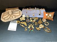 Full view of beaded purse, assorted brooches and Christmas themed pins, multiple with colored stones and gold tone metal, watches grouped on orange pillows