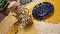 Photo showing the three-part china drip teapot with fruit design, small Belleek vessel with black tree motif, blue ceramic plate, and a selection of placemats underneath in parts of yellow and white
