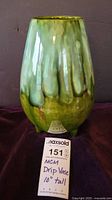 Front view of green ceramic footed vase with drip glaze on a dark fabric background, with lot tag visible.