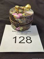 Front view of the small round porcelain box with floral design and gold bow on top, showing detailed clasp and artwork.