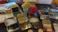 Wide photo showing multiple small metal tins filled with various screws, pins, cotton swabs, paper decals and small metal parts for model trains