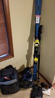Photo of K2 skis standing upright with Scott ski poles leaning against wall next to boots, boot bag, gloves, and goggles on floor.