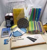 Photo showing assorted kitchenware including stove covers, straws, mitts, a sponge, trivet, peeler, spatula and packaged electric grinder.