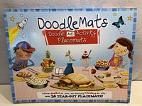 Front of box showing colorful placemat illustrations featuring children, food, a rocket, and butterflies. Title reads 'Doodle Mats Doodle and Activity Placemats' with note of 36 tear-out placemats.