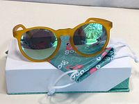 Yellow matte framed Goodr sunglasses with green mirrored lenses placed on top of a white box and teal Goodr pouch.