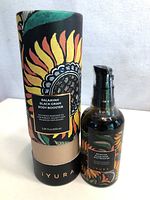 Photo of Iyura Balayayah Black Gram Body Booster massage oil 100ml bottle next to its decorated cardboard packaging tube, artwork includes sunflower and leaves in black, gold and green colors.