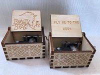 Photo showing two wooden music boxes with open lids, engraved with 'Beauty and the Beast' and 'Fly Me to the Moon'. Both boxes display visible mechanical music mechanisms inside and decorative patterns on the sides.