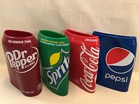 Four soda can shaped beer can covers in Dr Pepper, Sprite, Coca-Cola, and Pepsi branding and colors, displayed upright on a wooden surface.