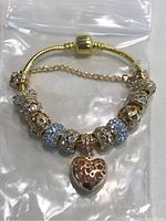 Gold-tone charm bracelet on snake chain with safety chain, multiple decorative beads including blue rhinestone beads and heart dangle charm.