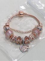 Rose gold plated S925 women's charm bracelet displayed on a white surface with protective plastic background, showing various pink and clear crystal adorned charms and heart detail with safety chain.