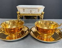 Two gold-tone antique Royal Winton Grimwades teacups with matching saucers, displayed with a footed ceramic soap dish in the background
