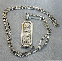 Silver chain necklace with decorative links and attached rectangular pendant with raised Egyptian hieroglyphic symbols on textured background.