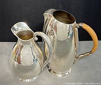 Two silver plated pitchers side by side on a gray surface under direct lighting showing polished reflective surfaces and wrapped handles.