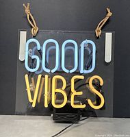 Good Vibes neon sign illuminated showing blue 'GOOD' and yellow 'VIBES' tubing mounted on clear acrylic with hanging ropes and power adapter visible.