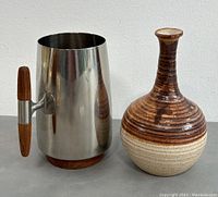 Stainless steel mug and signed pottery vase shown from front angle to display overall shape and materials.