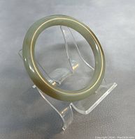 Jade bangle displayed on clear stand, showing smooth rounded surface and subtle color variations.