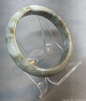 Natural jade bangle displayed on clear stand showing round shape and mixed green tones