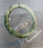 Natural jade bangle bracelet standing upright on clear plastic display stand showing gradient colors and polished finish.