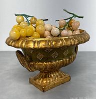 Gold-colored footed bowl with green diamond pattern filled with clusters of alabaster grapes in yellow and pink.