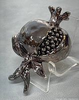 Front angled view of silver-coated pomegranate sculpture showing seeds and stem with leaves.