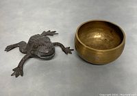 Photo showing vintage brass singing bowl and metal frog figurine side by side on flat surface