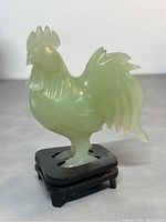Front and side view of light green carved jade rooster standing on a small carved wooden base, showing details of feathers and comb.