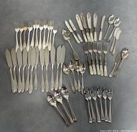 Full set spread out on grey surface, showing multiple types of cutlery: knives, forks, spoons, in silver plate with some featuring mother of pearl handles.