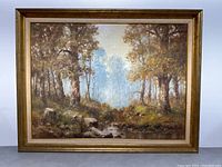 Framed landscape painting showing wooded forest scene, stream and rocks with visible artist signature in lower right corner
