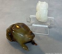 White jade Buddha figurine and greenish-brown stone frog figurine displayed side by side on a gray surface.