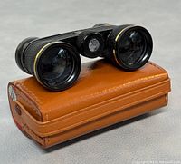 Vintage Milo opera binoculars resting on the original brown leather carrying case showing the coated lenses and textured black body with gold trim.