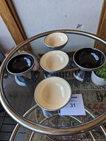 Five polished aluminum condiment containers on a glass table, three with beige/off white liners and two with black liners.