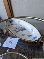 Angle view of blue and white floral oval serving dish with brass beaded rim and one handle visible, sitting on glass surface next to auction lot number.