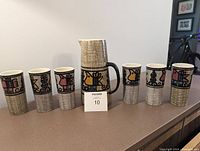 Front view showing pitcher centered with 6 tumblers arranged on either side, displaying the painted motifs and textured glaze.