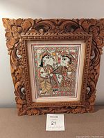 Wooden frame with intricate carving edges surrounding traditional Indian folk art print of two people in colorful attire facing each other.