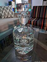 Full front view of clear crystal decanter with stopper on glass table, showing shape and thickness of the glass.