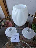 Three vintage glass shades displayed on a round glass table. One large white ribbed shade with an oval shape and two smaller frosted shades with concentric ridges.