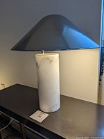 Full view of the table lamp showing the cylindrical white marble base and large conical polished metal shade, placed on a dark surface with lighting highlighting its texture and finish.