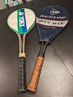 Pair of old school tennis rackets side by side; Yonex racket with green and white cover on the left, Dunlop Blue Max racket with blue cover on the right. Handles show black and brown grips respectively.