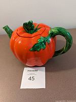 Front angle of red tomato-shaped ceramic teapot with green stem handle and leafy top lid on a table with lot number card