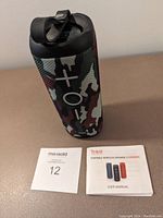 Tribit Stormbox speaker standing upright showing large plus, minus and power buttons in camouflage fabric with user manual and lot number card on surface.