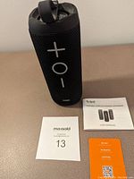 Black Tribit Stormbox Bluetooth speaker showing front with volume control (+, o, -) and loop strap, next to user manual and warranty card on beige surface.